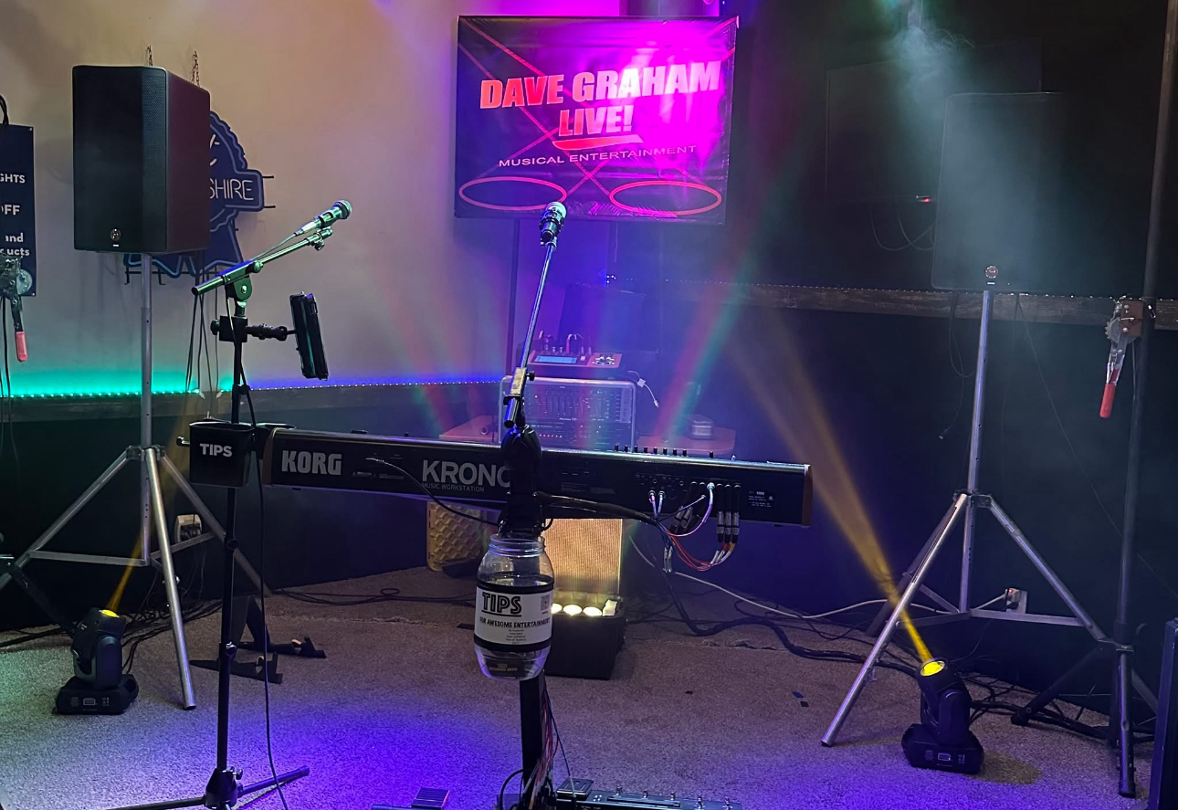 Dave Graham performing live at the keyboard with stage lights, banner, and professional audio equipment.