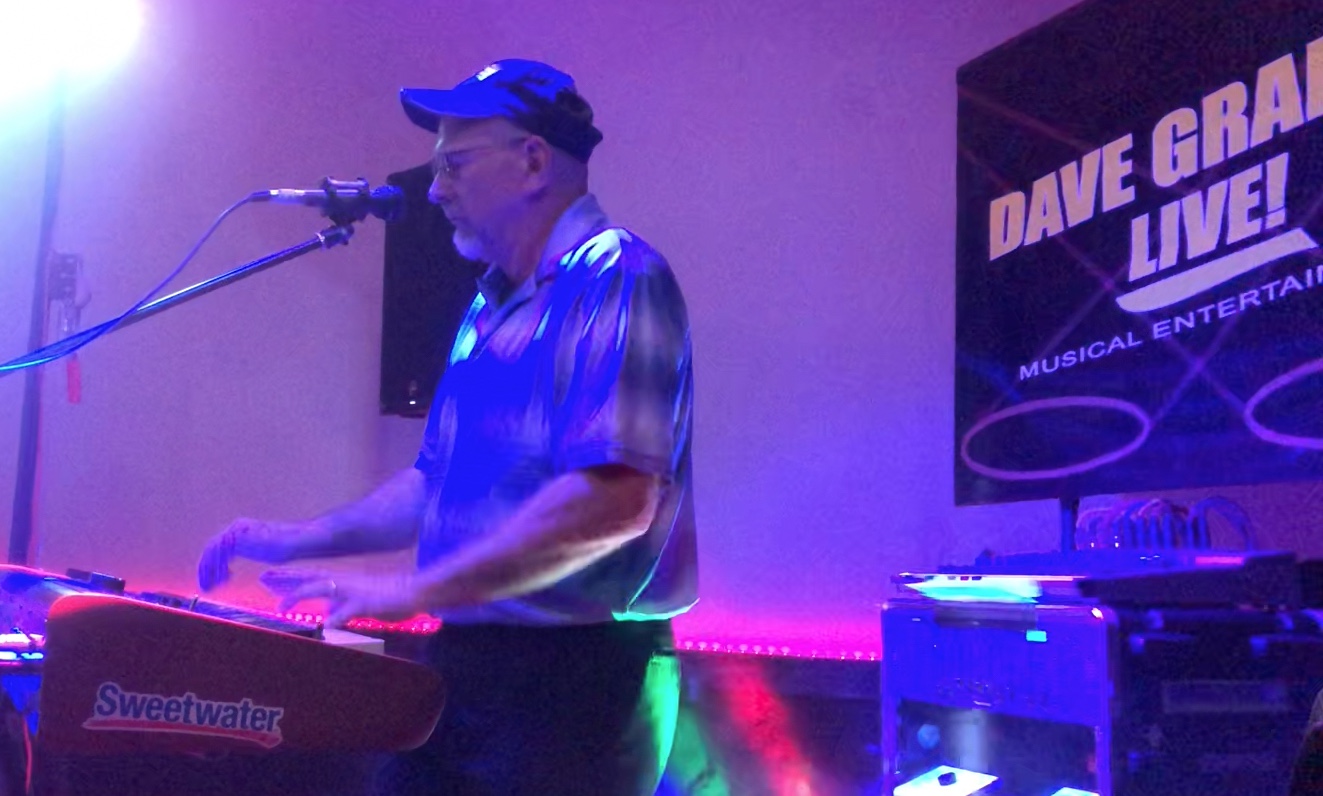 Dave Graham Live performance rig setup in a bright indoor space with a Korg Kronos keyboard, electric guitar, stage lighting, and a branded t-shirt on display.